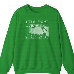 Peut inclure: Un sweat-shirt vert avec l'inscription blanche "TITLE FIGHT". Le motif repr&eacute;sente une personne tombant d'un h&eacute;licopt&egrave;re, avec une foule en dessous. Le sweat-shirt est fait d'une mati&egrave;re douce.