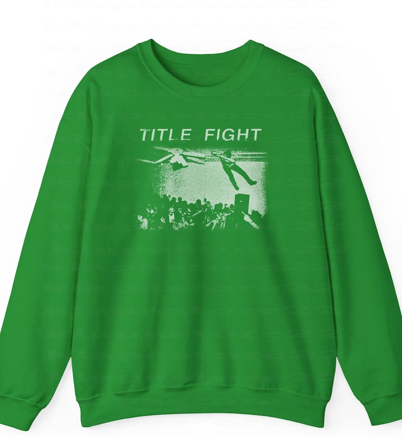 Peut inclure: Un sweat-shirt vert avec l'inscription blanche "TITLE FIGHT". Le motif repr&eacute;sente une personne tombant d'un h&eacute;licopt&egrave;re, avec une foule en dessous. Le sweat-shirt est fait d'une mati&egrave;re douce.