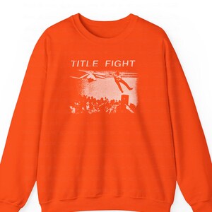Peut inclure: Sweat-shirt orange vif avec l'inscription "TITLE FIGHT" au-dessus d'un motif blanc repr&eacute;sentant une personne tombant d'une sc&egrave;ne, avec une foule en dessous. Le sweat-shirt est de style col rond.