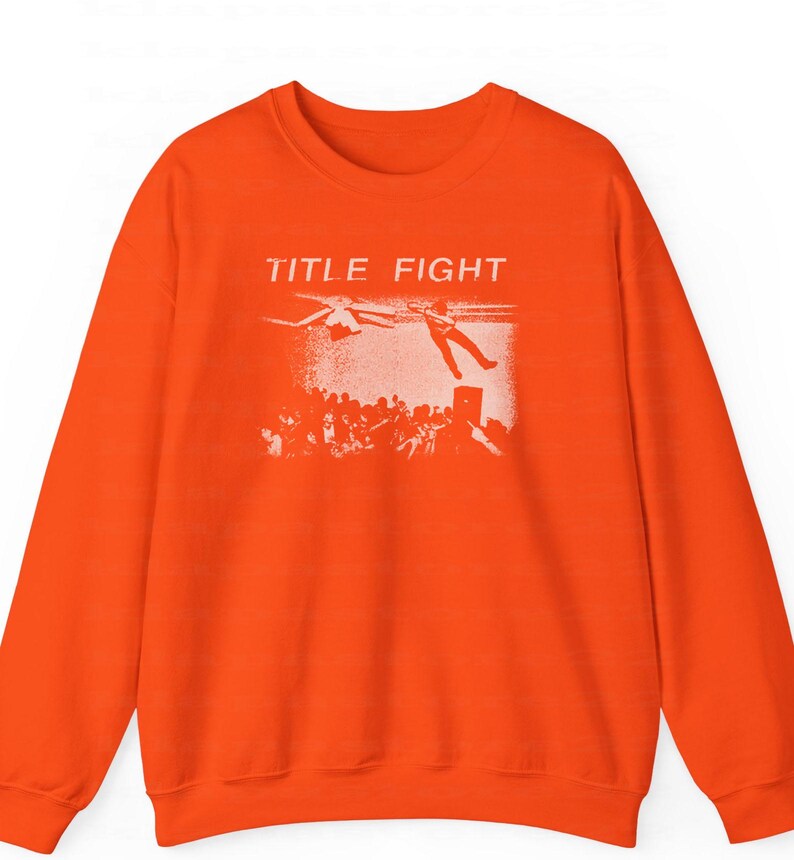 Peut inclure: Sweat-shirt orange vif avec l'inscription "TITLE FIGHT" au-dessus d'un motif blanc repr&eacute;sentant une personne tombant d'une sc&egrave;ne, avec une foule en dessous. Le sweat-shirt est de style col rond.