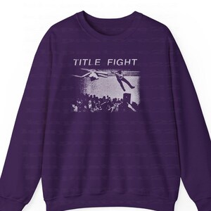 Peut inclure: Un sweat-shirt violet fonc&eacute; avec un graphisme blanc repr&eacute;sentant une sc&egrave;ne de concert et le texte "TITLE FIGHT". Le graphisme montre une personne tombant d'une sc&egrave;ne avec une foule en dessous. Le sweat-shirt a des manches longues et un col rond.