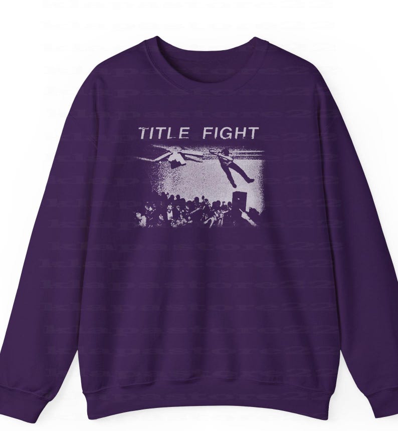 Peut inclure: Un sweat-shirt violet fonc&eacute; avec un graphisme blanc repr&eacute;sentant une sc&egrave;ne de concert et le texte "TITLE FIGHT". Le graphisme montre une personne tombant d'une sc&egrave;ne avec une foule en dessous. Le sweat-shirt a des manches longues et un col rond.