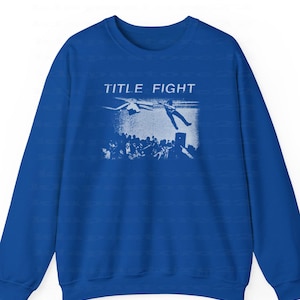 Peut inclure: Sweat-shirt bleu roi avec un motif blanc. Le motif affiche le texte "TITLE FIGHT" et une personne tombant d'une sc&egrave;ne, avec une foule en dessous. Le sweat-shirt a des manches longues et un col rond.