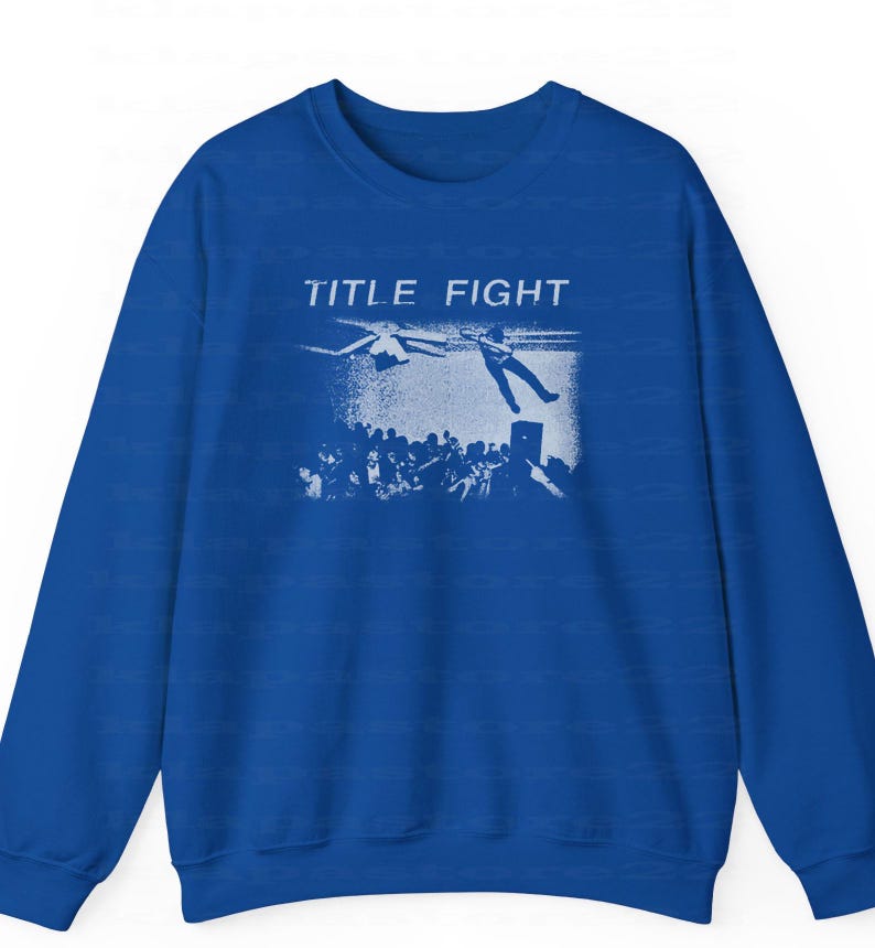 Peut inclure: Sweat-shirt bleu roi avec un motif blanc. Le motif affiche le texte "TITLE FIGHT" et une personne tombant d'une sc&egrave;ne, avec une foule en dessous. Le sweat-shirt a des manches longues et un col rond.