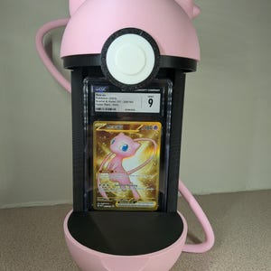 May include: A pink 3D-printed Pok&eacute;mon card holder shaped like Mew, holding a graded Mew ex card. The card is in a protective case, featuring a gold and pink Mew illustration. The holder has a black interior and a circular design.