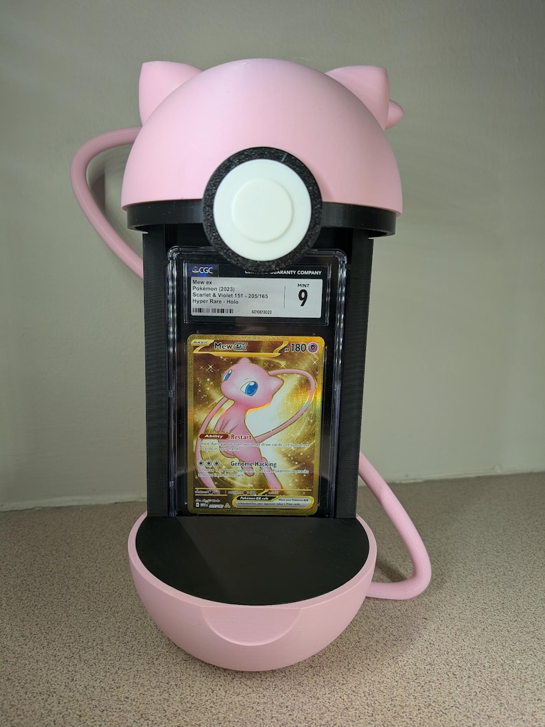 May include: A pink 3D-printed Pok&eacute;mon card holder shaped like Mew, holding a graded Mew ex card. The card is in a protective case, featuring a gold and pink Mew illustration. The holder has a black interior and a circular design.