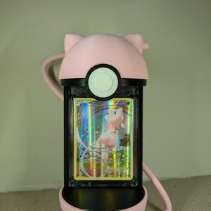 May include: A pink, 3D-printed Pok&eacute; Ball-shaped card holder with a black frame. The holder contains a holographic Pok&eacute;mon card featuring Mew. The holder has a loop for carrying and a cat-ear design on top.