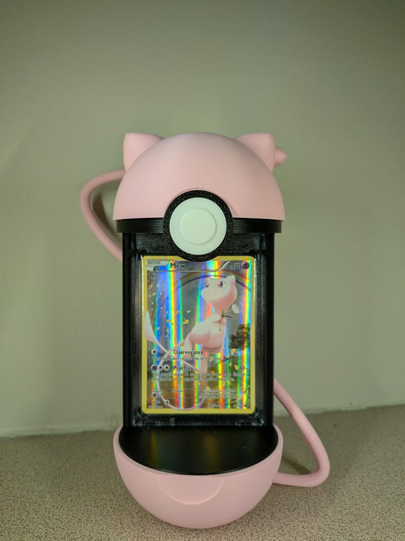 May include: A pink, 3D-printed Pok&eacute; Ball-shaped card holder with a black frame. The holder contains a holographic Pok&eacute;mon card featuring Mew. The holder has a loop for carrying and a cat-ear design on top.