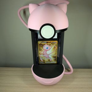 May include: A pink and black display case shaped like a Pok&eacute; Ball, with a Mew Pok&eacute;mon card inside. The case has a rounded top with cat-like ears and a circular opening. The card features a golden background and a pink Mew character.