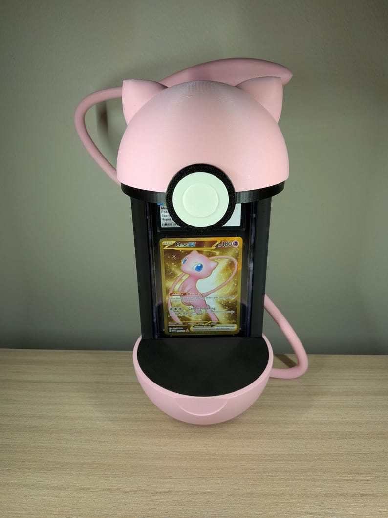 May include: A pink and black display case shaped like a Pok&eacute; Ball, with a Mew Pok&eacute;mon card inside. The case has a rounded top with cat-like ears and a circular opening. The card features a golden background and a pink Mew character.