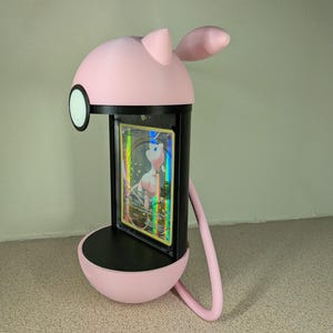 May include: A pink and black card display stand shaped like a cartoon character. The stand holds a trading card with a holographic design. The stand has a round base, a cylindrical body, and a head with ears. A curved arm extends from the head to the base.