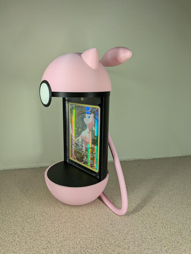 May include: A pink and black card display stand shaped like a cartoon character. The stand holds a trading card with a holographic design. The stand has a round base, a cylindrical body, and a head with ears. A curved arm extends from the head to the base.