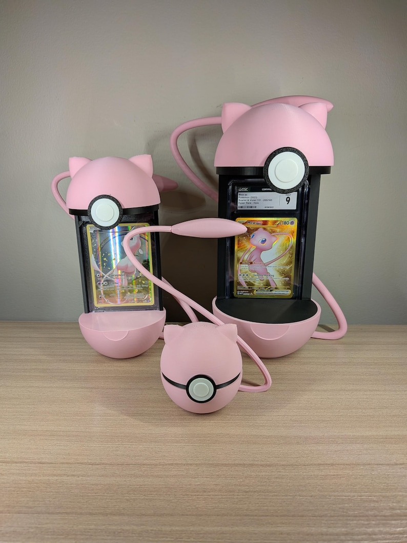 May include: Pink and black card holders in the shape of a cartoon character. The holders have a circular base and a top with ears. A small pink ball with a black stripe and white circle is also present. The holders contain trading cards.