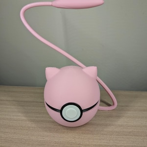 May include: A pink desk lamp with a rounded base and a curved, flexible neck. The base is spherical with black accents and a white button. The lamp head is oval-shaped. The lamp is on a wooden surface.
