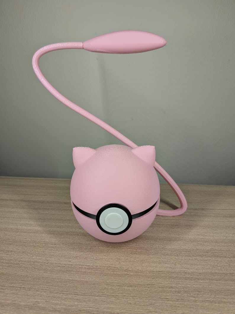 May include: A pink desk lamp with a rounded base and a curved, flexible neck. The base is spherical with black accents and a white button. The lamp head is oval-shaped. The lamp is on a wooden surface.