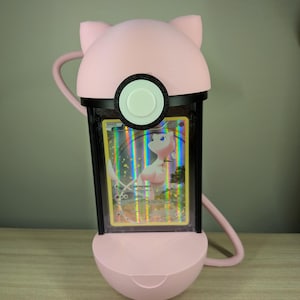 May include: A pink, 3D-printed Pok&eacute;mon card holder shaped like a character's head, with ears and a circular button. The holder encases a holographic card featuring a pink Pok&eacute;mon. The holder has a black frame and a curved handle.