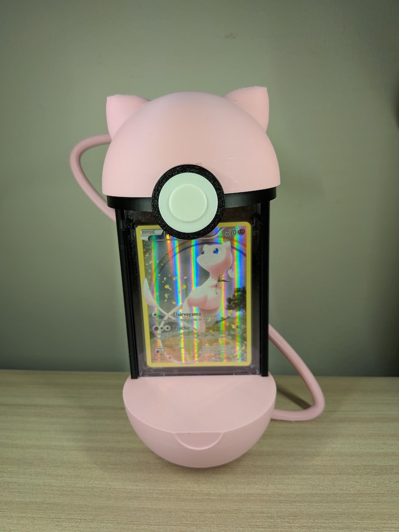 May include: A pink, 3D-printed Pok&eacute;mon card holder shaped like a character's head, with ears and a circular button. The holder encases a holographic card featuring a pink Pok&eacute;mon. The holder has a black frame and a curved handle.