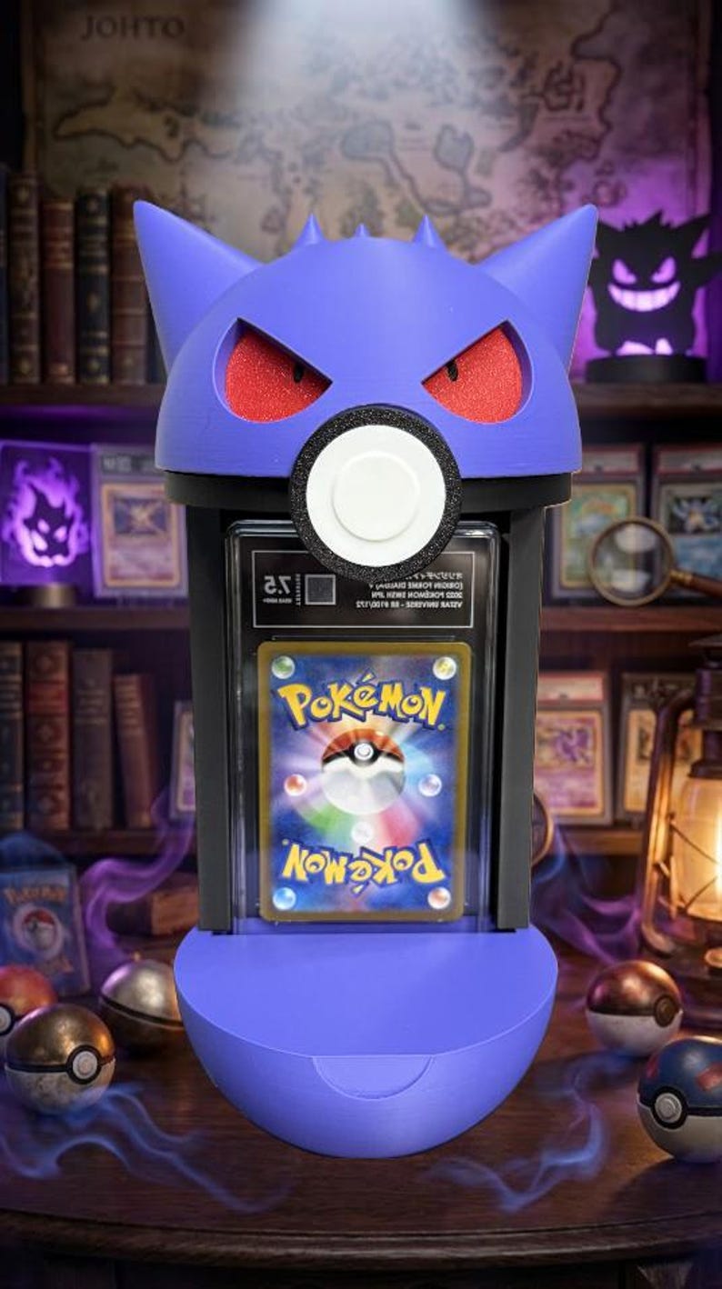 Pode incluir: Um suporte de cart&atilde;o roxo com tema Gengar com olhos vermelhos e um c&iacute;rculo branco. O suporte exibe um cart&atilde;o Pok&eacute;mon. O fundo inclui uma estante, outros itens com tema Pok&eacute;mon e a palavra "JOHTO".