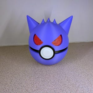 Pode incluir: Uma esfera roxa impressa em 3D com uma faixa preta e um c&iacute;rculo branco, assemelhando-se a uma Pok&eacute; Ball. O topo apresenta orelhas pontudas e espinhos, com cavidades oculares vermelhas texturizadas. Um colecion&aacute;vel &uacute;nico e estilizado.