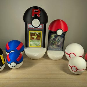 May include: A collection of spherical containers resembling Pokeballs in various colors including red, white, blue, black, and yellow. Two containers are designed as vertical display stands holding rectangular trading cards, one featuring a red R logo.