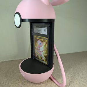 May include: A pink and black display stand shaped like a cartoon character, holding a graded trading card. The stand has a round base, a rectangular card holder, and a head with ears. The card features a gold and pink illustration.