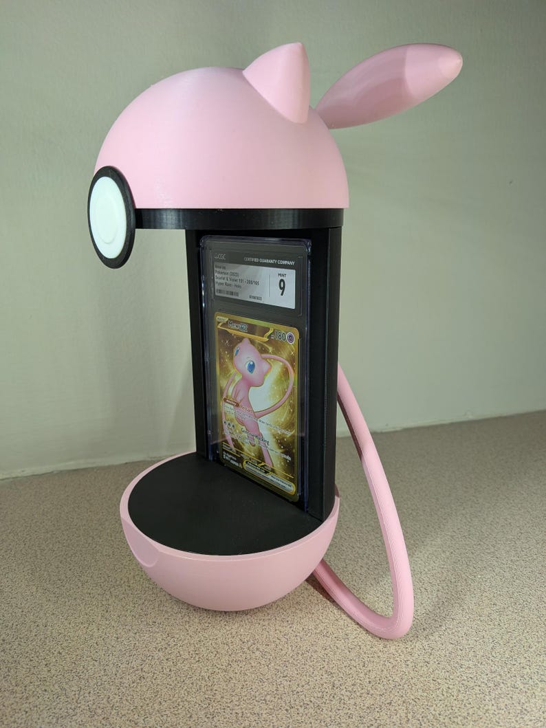 May include: A pink and black display stand shaped like a cartoon character, holding a graded trading card. The stand has a round base, a rectangular card holder, and a head with ears. The card features a gold and pink illustration.