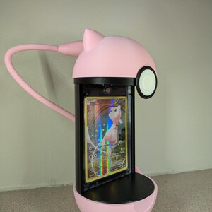 May include: A pink, cat-shaped desk lamp with a flexible arm, illuminating a Pokemon trading card. The card is encased in a protective holder, featuring a holographic design with a pink and white creature.