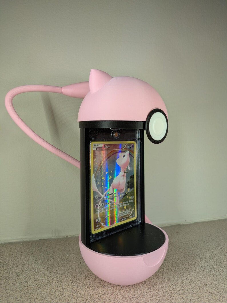 May include: A pink, cat-shaped desk lamp with a flexible arm, illuminating a Pokemon trading card. The card is encased in a protective holder, featuring a holographic design with a pink and white creature.