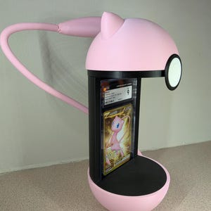 May include: A pink desk lamp with a black base and a flexible arm. The lamp has a circular head with a white and black accent. A graded trading card featuring a pink Pokemon is displayed in the lamp's center.