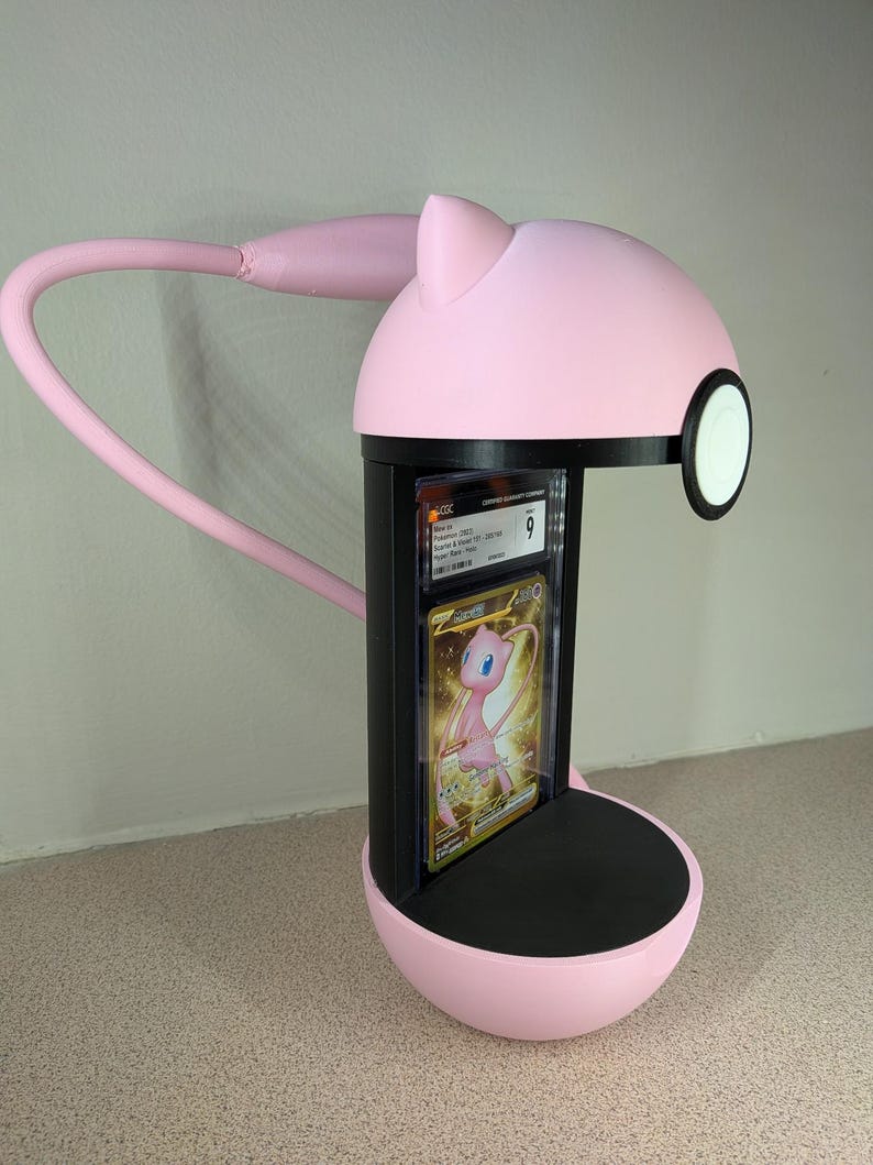 May include: A pink desk lamp with a black base and a flexible arm. The lamp has a circular head with a white and black accent. A graded trading card featuring a pink Pokemon is displayed in the lamp's center.