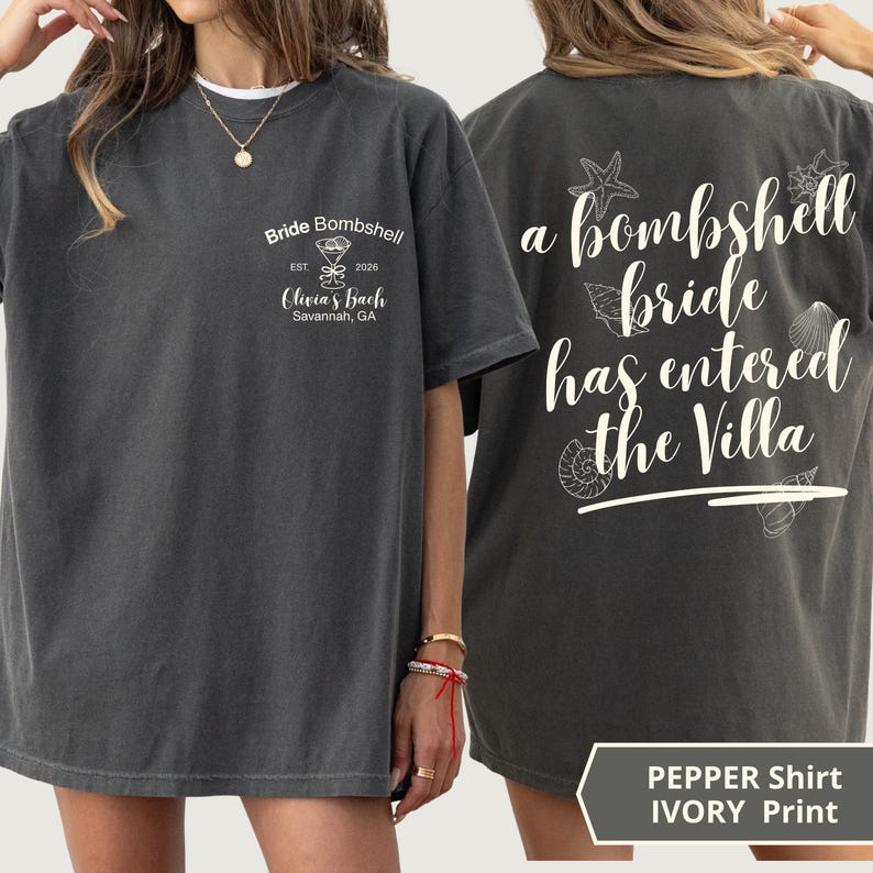 K&ouml;nnte beinhalten: Ein anthrazitfarbenes T-Shirt mit wei&szlig;em Text und Grafiken. Die Vorderseite zeigt ein Martiniglas-Symbol mit dem Text Bride Bombshell, Est 2026, Olivia's Bach, Savannah, GA. Die R&uuml;ckseite zeigt den Text a bombshell bride has entered the Villa.