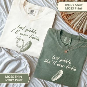 Peut inclure: Deux t-shirts ivoire et vert mousse avec les phrases "last pickle I'll ever tickle" et "last pickle she'll ever tickle" imprimées. Chaque chemise a un motif de cornichon et de bague.