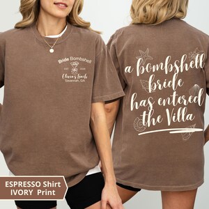 K&ouml;nnte beinhalten: Ein espressofarbenes Kurzarm-T-Shirt mit elfenbeinfarbenem Text und Grafiken. Die Vorderseite zeigt ein Martiniglas-Symbol mit dem Text Bride Bombshell, Est. 2026, Olivia's Bach, Savannah, GA. Die R&uuml;ckseite zeigt den Text a bombshell bride has entered the Villa mit Muschelillustrationen.