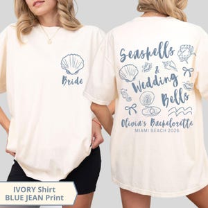 May include: Cream-coloured t-shirt with a blue jean print. The front has a seashell graphic and the word "Bride." The back displays "Seashells & Wedding Bells," "Olivia's Bachelorette," and "Miami Beach 2026" with seashell and nautical illustrations.