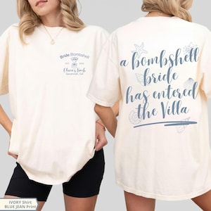 K&ouml;nnte beinhalten: Ein elfenbeinfarbenes T-Shirt mit blauem Text und Grafiken. Die Vorderseite zeigt "Bride Bombshell EST 2026 Olivia's Bach Savannah, GA" mit einem Martiniglas. Die R&uuml;ckseite zeigt "a bombshell bride has entered the Villa" mit Muschelgrafiken.