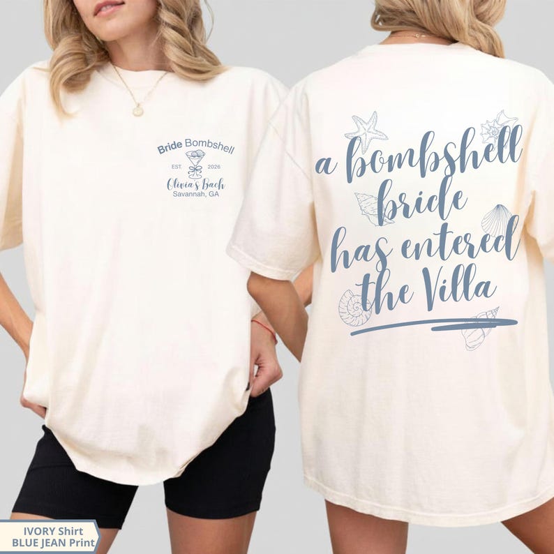 K&ouml;nnte beinhalten: Ein elfenbeinfarbenes T-Shirt mit blauem Text und Grafiken. Die Vorderseite zeigt "Bride Bombshell EST 2026 Olivia's Bach Savannah, GA" mit einem Martiniglas. Die R&uuml;ckseite zeigt "a bombshell bride has entered the Villa" mit Muschelgrafiken.