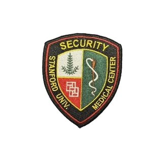 Puede incluir: Parche bordado con diseño de escudo. El parche presenta las palabras "SECURITY", "STANFORD UNIV." y "MEDICAL CENTER". El escudo está dividido en secciones con un árbol, un símbolo médico y un diseño geométrico. El parche tiene un borde negro.