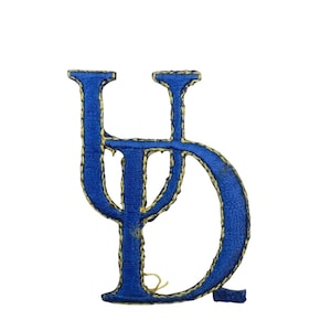 May include: Embroidered blue letter patch with a gold border. The patch features a stylized design of the letters "LD" in a connected script. The patch is likely for clothing or accessories.