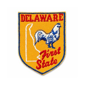Pode incluir: Patch bordado com o estado de Delaware. O patch tem um fundo amarelo com uma borda azul. Inclui um contorno branco do estado, um galo e as palavras "DELAWARE First State" em vermelho.