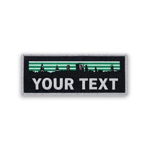 Pode incluir: Remendo retangular preto com borda prateada. O remendo apresenta um design listrado verde e branco acima do texto "YOUR TEXT" em letras maiúsculas brancas. Uma silhueta de uma paisagem urbana é visível dentro das listras verdes.