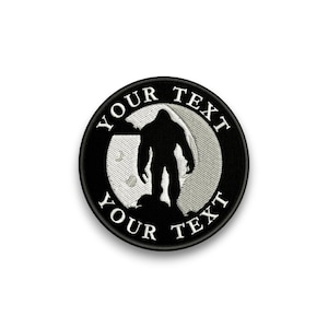Op de afbeelding: Een ronde, zwart geborduurde patch met een silhouet van een Bigfoot-wezen voor een getextureerde, lichtgrijze cirkel. De woorden "YOUR TEXT" zijn in het wit rond de cirkel geborduurd.