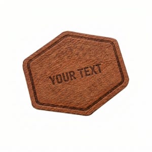 Puede incluir: Un parche de cuero marrón con una forma hexagonal única. El parche tiene una superficie texturizada y un borde marrón más oscuro. Las palabras "YOUR TEXT" están grabadas en el centro, ideal para la personalización.