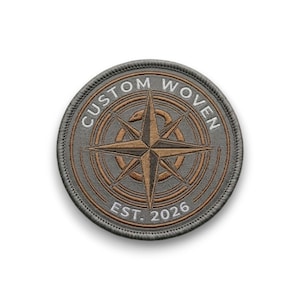Op de afbeelding: Een ronde, op maat geweven patch met een kompasroosontwerp in bruin- en grijstinten. De tekst "CUSTOM WOVEN" loopt in een boog over de bovenkant en "EST. 2026" staat onderaan. De patch heeft een gestikte rand.