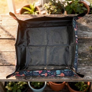 May include: A dark blue gardening tray with a black interior, decorated with a pattern of mushrooms and flowers. The tray is open and placed on a wooden surface, suitable for potting plants. Dimensions are not visible.