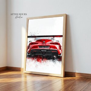 May include: A framed art print showing the rear view of a red sports car, rendered in a watercolour style with red and black paint splashes. The print is in a light wood frame, leaning against a white wall. The text "AFTER HOURS studio" is visible.