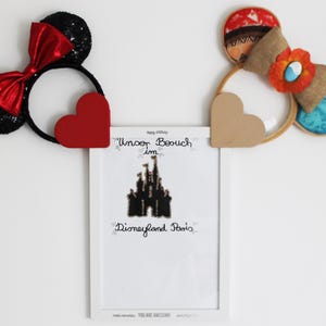 Könnte beinhalten: Zwei dekorative Stirnbänder mit Mickey Mouse Ohren, eines schwarz mit roten Pailletten und einer roten Schleife, das andere mit einer braunen Schleife und floralen Akzenten. Ein gerahmter Druck mit dem Text "Unser Besuch im Disneyland Paris" ist ebenfalls sichtbar.