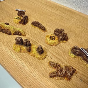 Puede incluir: Una colección de piezas de terreno en miniatura, con una variedad de formaciones rocosas en tonos marrones y amarillos. Algunas piezas tienen etiquetas de texto, incluyendo "HABBANYA RIDGE" y otra con un símbolo. Estas piezas son probablemente para juegos de mesa o modelismo.