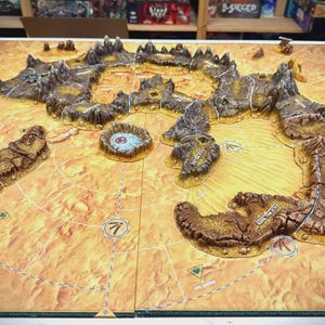 Könnte beinhalten: Ein detailliertes Brettspiel-Landschaft mit erhöhten Geländemerkmalen, darunter Berge und ein zentraler Krater. Das Brett hat ein beige-braunes Farbschema mit einem Rastermuster und nummerierten Markierungen. Das Spielbrett ist für strategisches Gameplay konzipiert.