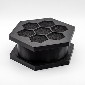 Può includere: Filtro dell'aria esagonale nero con un design a nido d'ape sulla parte superiore. Il filtro è composto da una base, una sezione centrale cilindrica e un coperchio con sei aperture esagonali riempite con un materiale scuro e poroso. Design moderno e geometrico.