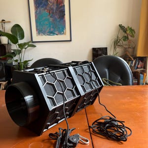 May include: A black rectangular device with a cylindrical opening on one side and a honeycomb pattern on the front. Black cords with adjustable clips extend from the device. The device is on a wooden table.